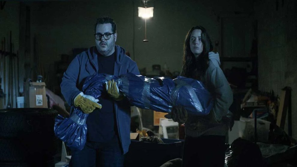 Kaya Scodelario and Josh Gad standing in a garage holding a body wrapped in plastic in a still from Alex Winter's "Aduthood."